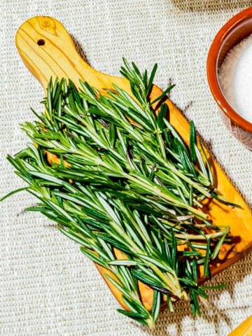 Ingredients for rosemary salt - rosemary sprigs on a small cutting board next to a brown enamel cup of salt