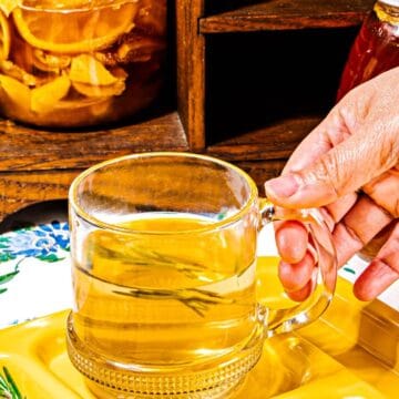 person holding handle of clear glass mug with pile of honey lemons and sprig of rosemary in foreground