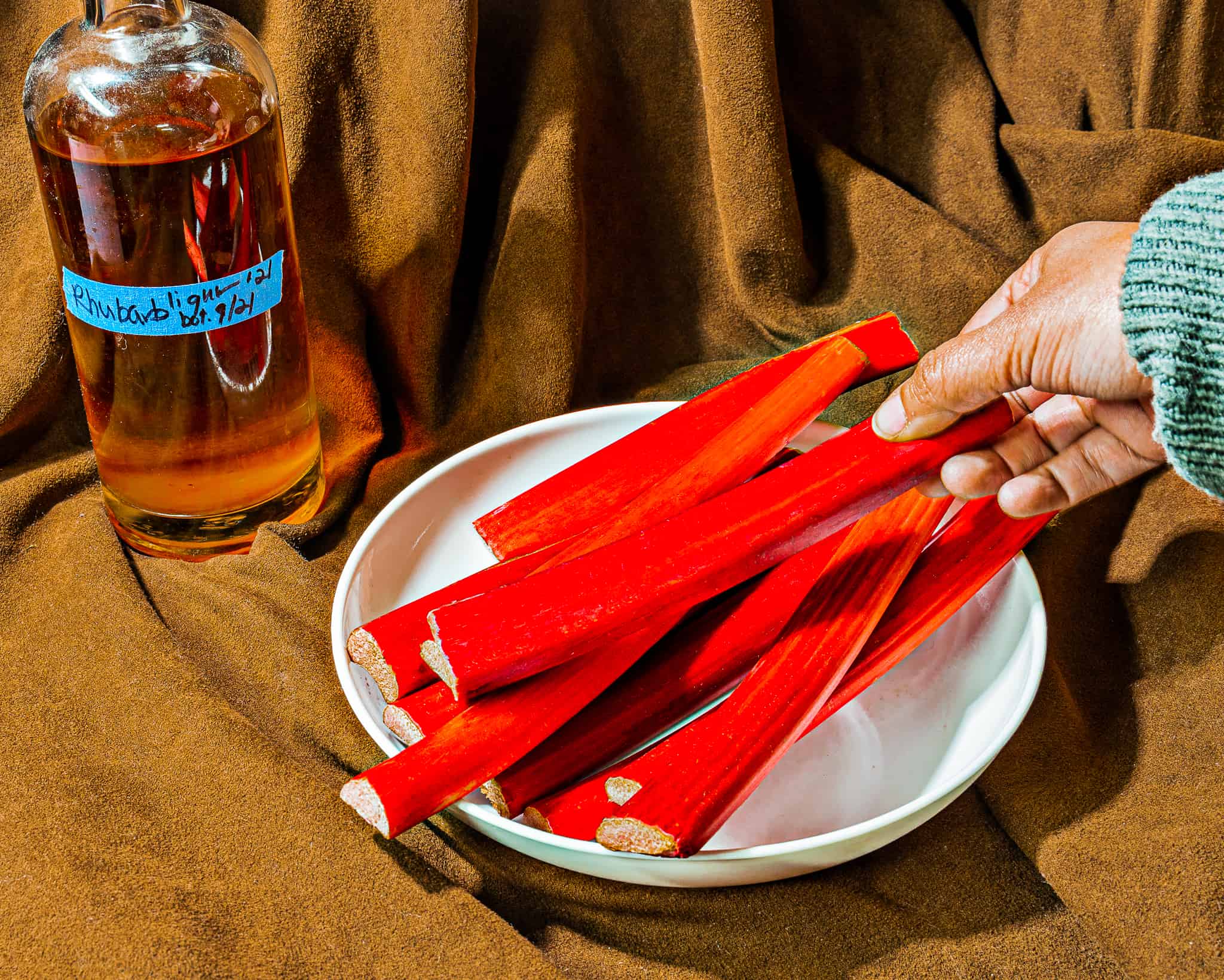 person picking up rhubarb stalk from a white bowl with a bottle of rhubarb liqueur in background