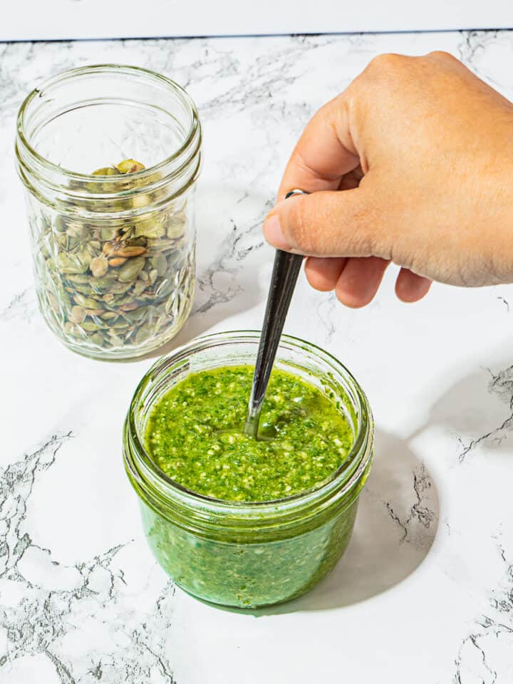 person pulling spoon out of garlic mustard pesto sauce with pepitas on a white background