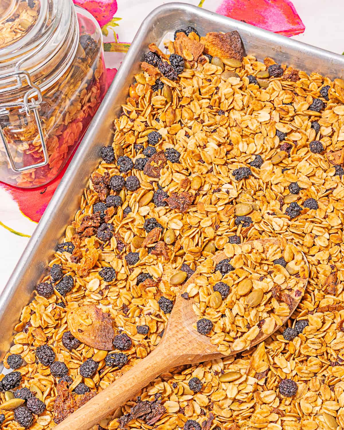 granola on a metal sheet tray on a pink and white patterned table cloth with a small glass jar full of granola 