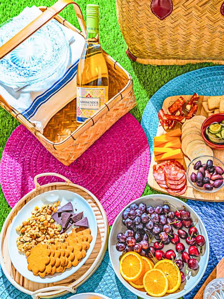 picnic setting with two picnic baskets, bottle of wine, charcuterie board, two plates of fruit and snacks