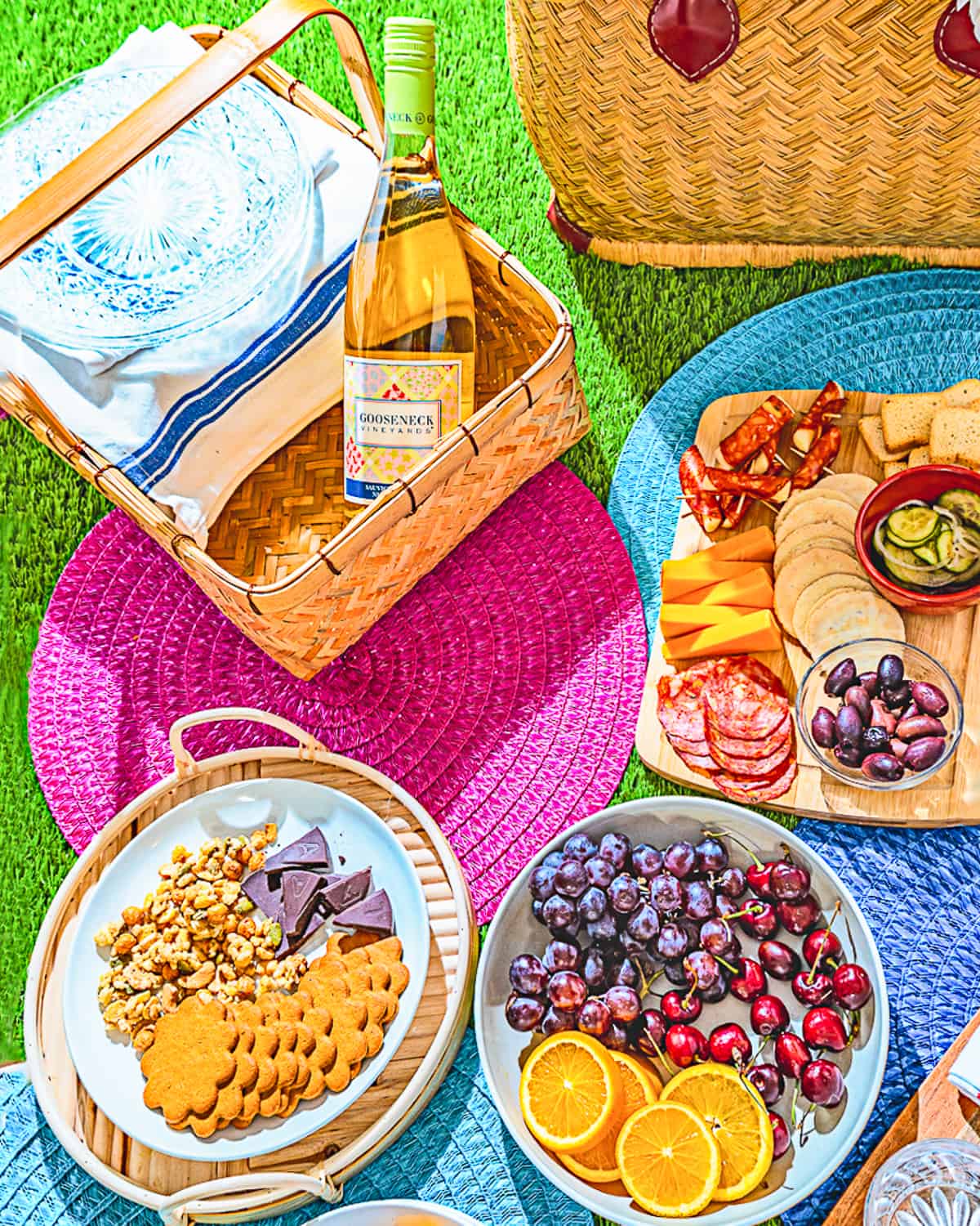 picnic setting with two picnic baskets, bottle of wine, charcuterie board, two plates of fruit and snacks