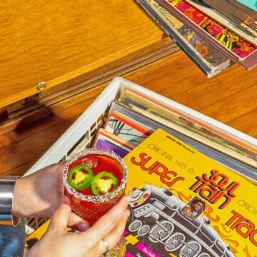 person holding pink jalapeno margarita while looking at a milk crate of records