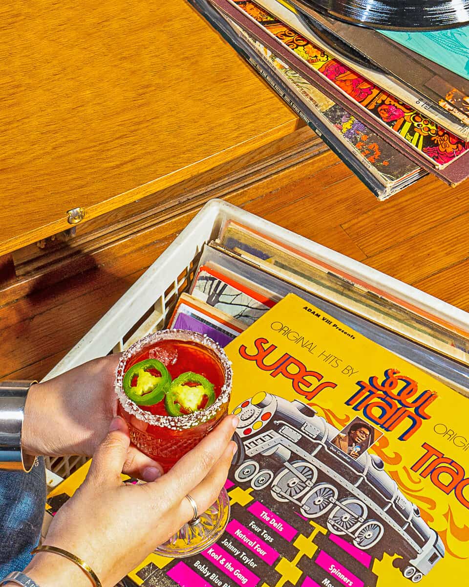 person holding pink jalapeno margarita while looking at a milk crate of records