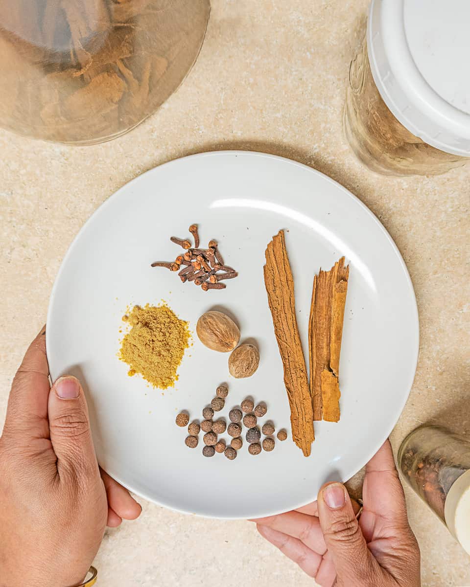 person holding a place of whole cloves, cinnamon bark, ground ginger, whole nutmeg, and allspice on a white plate