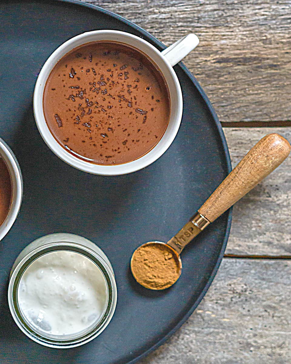 dairy free hot chocolate in a white mug, a measuring spoon with ground cinnamon, and pot of cream on a black plate on a wooden table