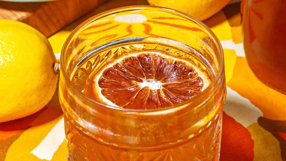 Glass of brown liquid (honey old fashion cocktail), garnished with dehydrated orange slice lemons, honey and a jar of lemon fermented honey in the background