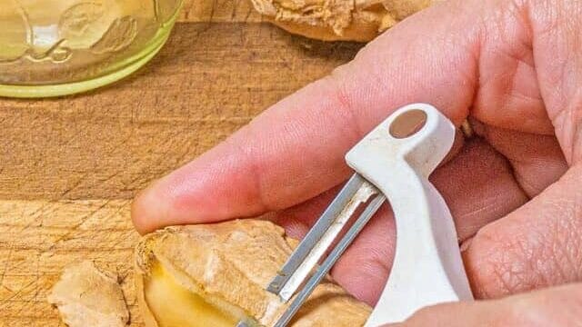 person peeling ginger with a vegetable peeler on a wooden cutting board