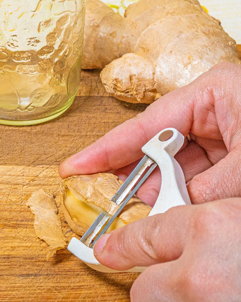 person peeling ginger with a vegetable peeler on a wooden cutting board