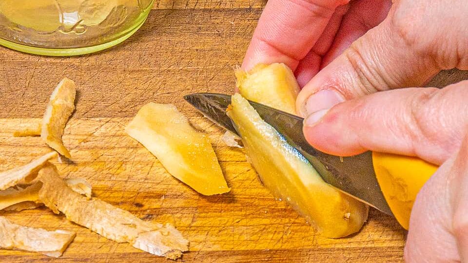 person slicing ginger with a pairing knife on a wooden cutting board