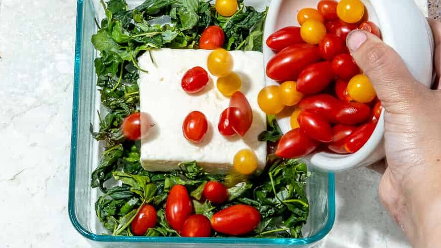 person adding tomatoes to baking dish of feta and greens