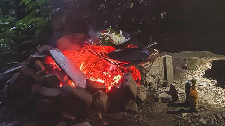 person shining light over open fire with cooking grate and cast iron skillet in the woods